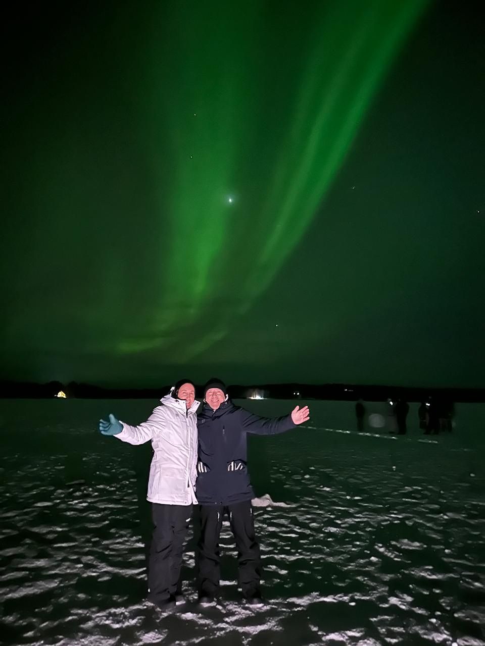 Two guests with arms open wide on a frozen lake under aurora rays