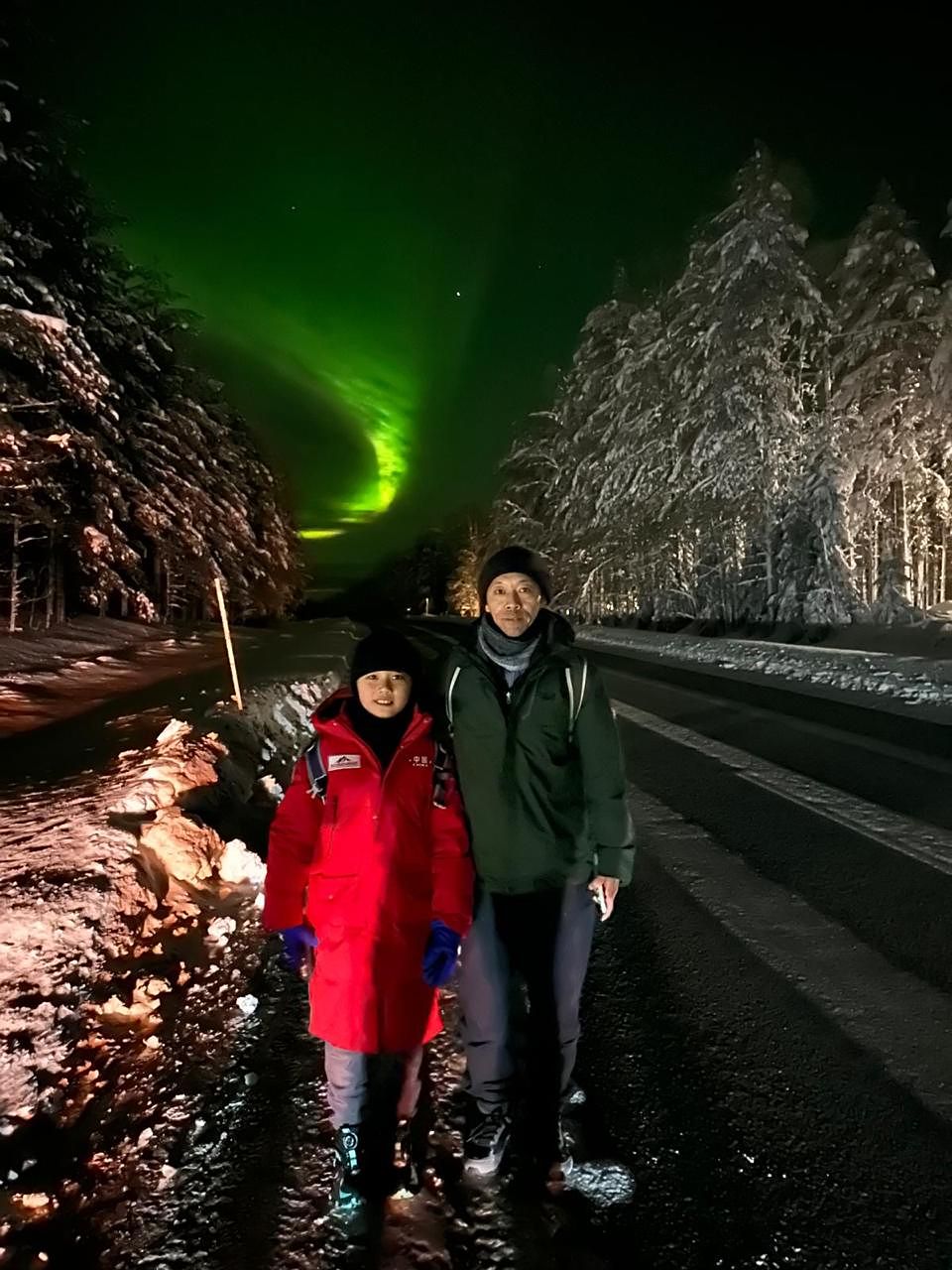 Two guests on a quiet road lined with snow-laden trees, aurora glowing at the end of it