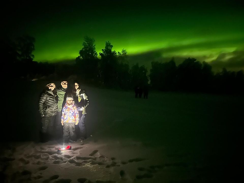 A family on a snowfield with a bright green aurora along the horizon