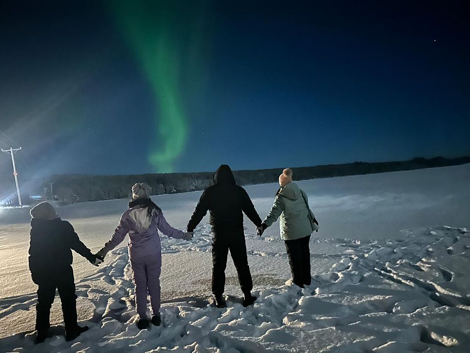 Four guests holding hands on a moonlit frozen lake, looking up at the aurora