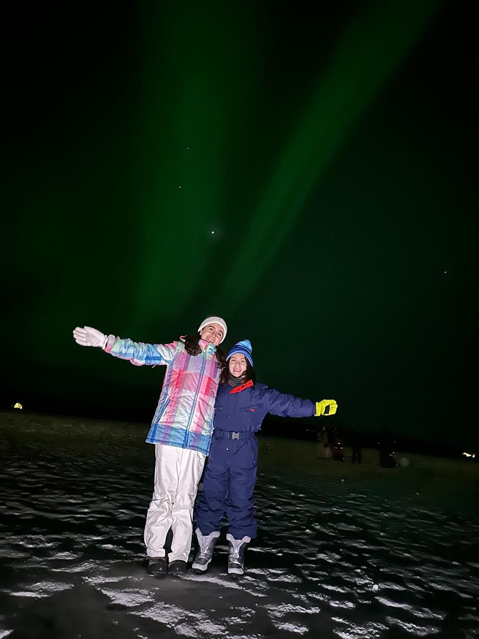 Two friends in ski wear under tall green aurora rays