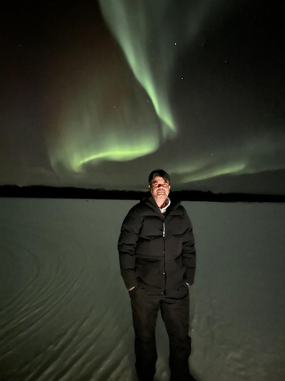 A guest standing on a frozen lake as the aurora curls overhead