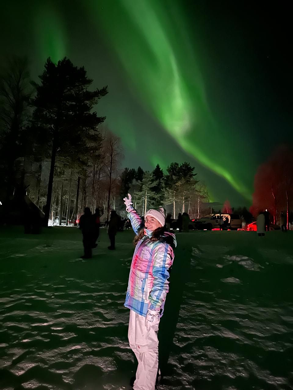 A guest reaching up towards a sweeping aurora band by a pine forest