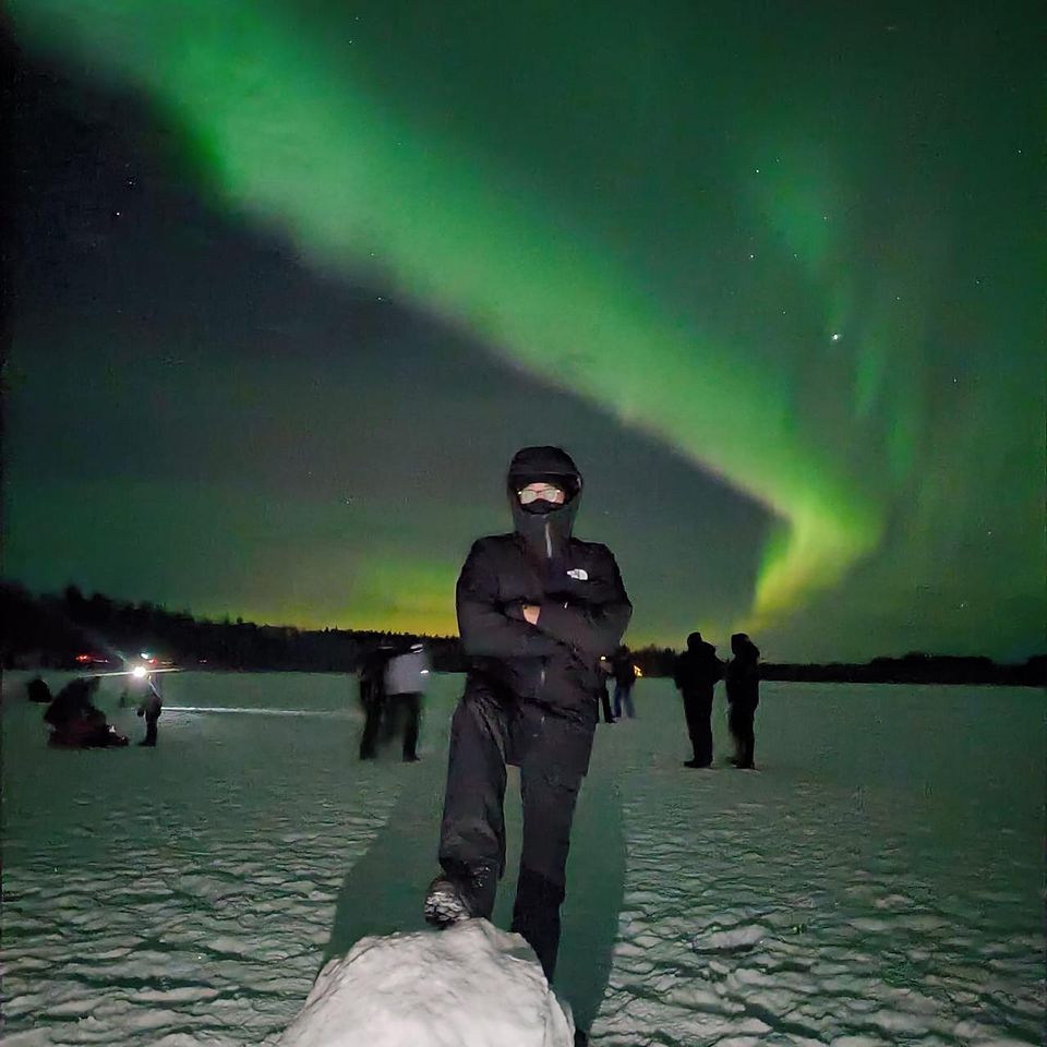 A guest on a snow mound beneath a wide aurora arc over a frozen lake
