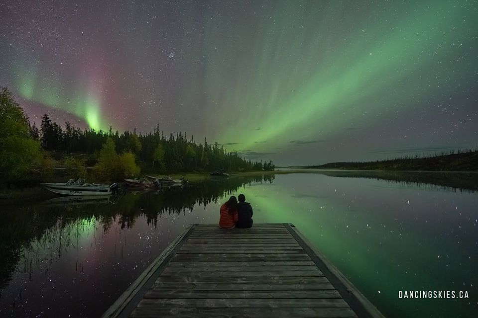 A couple sitting at the end of a wooden dock as the aurora reflects in a still lake