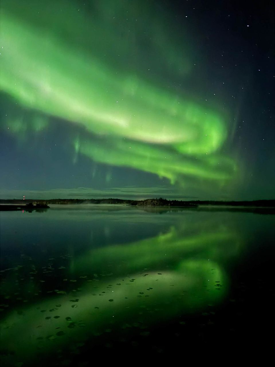 A coiled green aurora band above a calm lake dotted with lily pads