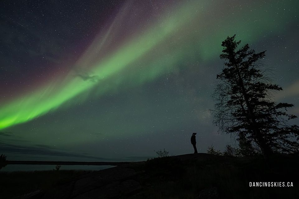 A lone figure on a rocky rise beneath a broad green aurora arc