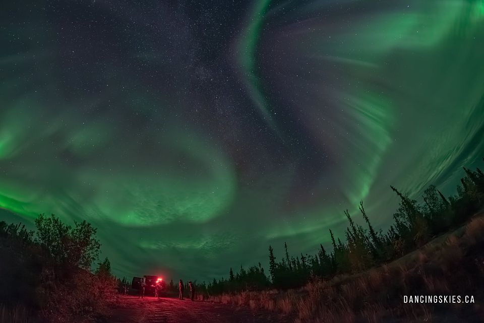 A wide aurora swirl over a forest clearing, a vehicle’s red tail lights below