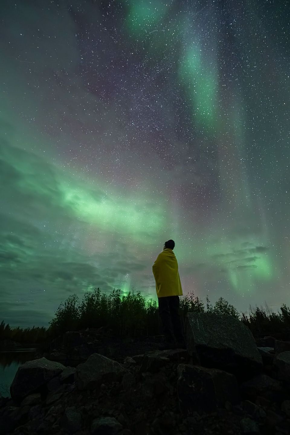 A person in a yellow jacket standing on rocks, looking up at green aurora and stars